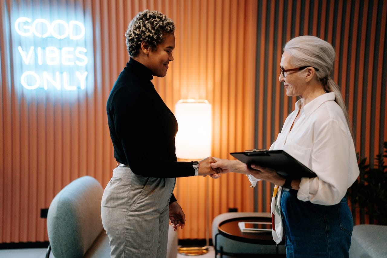 one woman shaking hands with another woman after recruiting her. Neon sign about good vibes in the background of their office.
